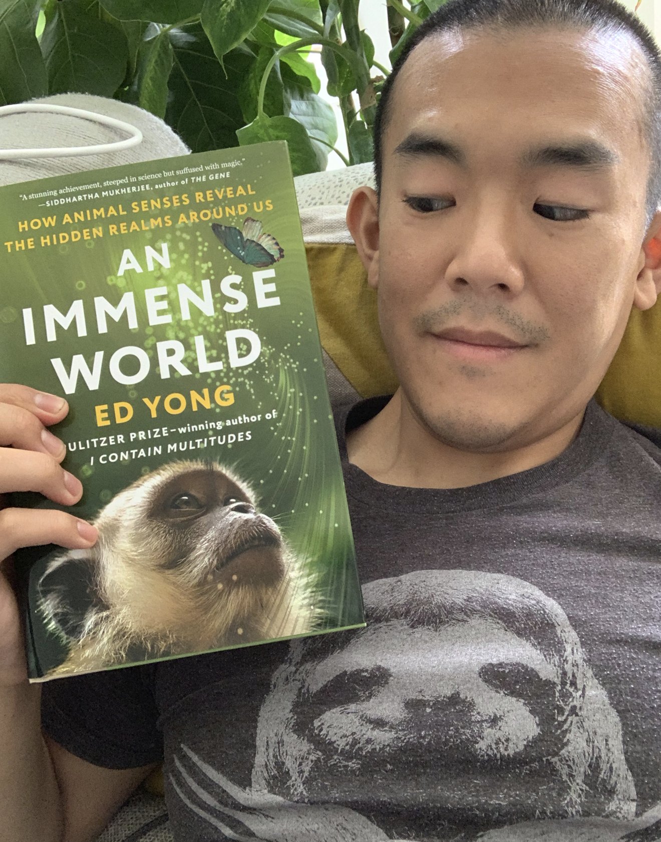 Selfie of author Ed Yong looking at the cover of his book An Immense World, which features a blue butterfly and a primate's face over a green background.