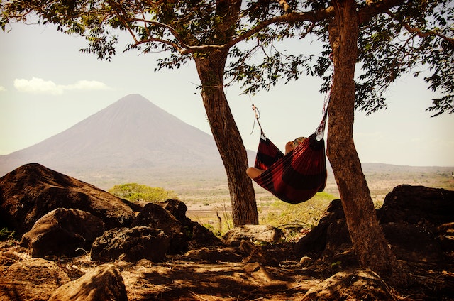 A dark red hammock suspended between two trees, overlooking a flat stretch of desert and a mountain in the distance. Photo by Leonie Fahjen via Pexels.