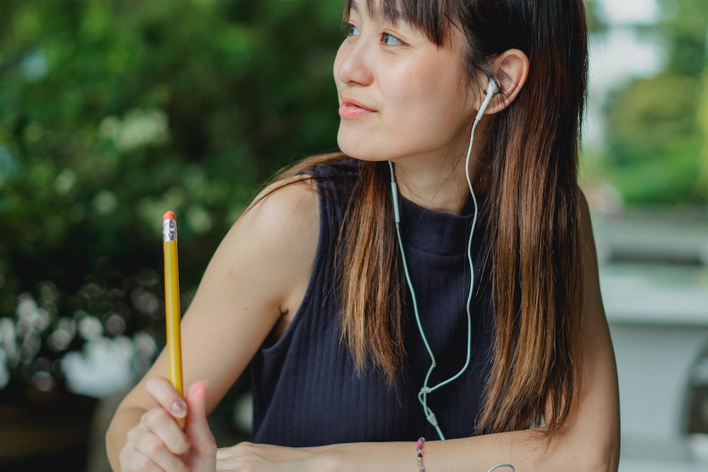 Woman of Asian descent in a navy tank top with long brown hair looking to her right, wearing earbuds, and holding a pencil upright in her right hand. Photo by Zen Chung via Pexels.