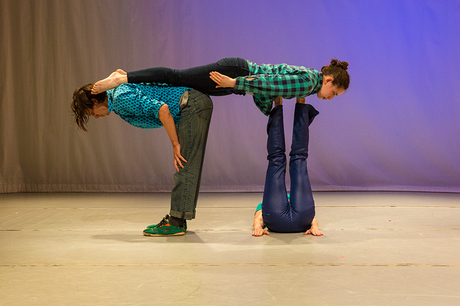 A male person in jeans and a turquoise shirt standing and bent forward at the waist at a 90 degree angle. Next to him is a woman lying on her back with her legs in the air. Another woman is horizontal with her chest on the woman's feet and her legs on the man's back.