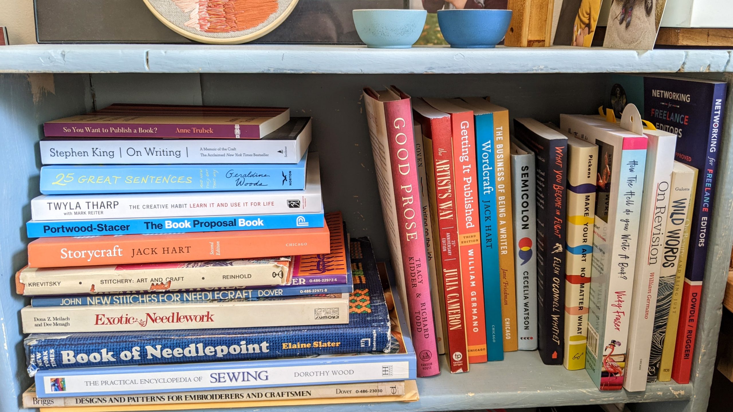 A pale blue, painted bookshelf. Some books on embroidery and needlepoint are stacked on their side with some writing craft books on top, and more writing and editing books are upright next to them. On top of the bookshelf, the bottom edge of an embroidery piece, two tiny wooden bowls, and several photographs are visible.