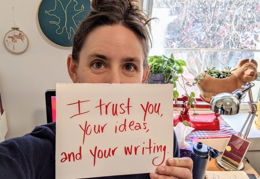 A white woman with curly brown hair in a bun and hazel eyes holds a sign in front the bottom half of her face saying "I trust you, your ideas, and your writing."