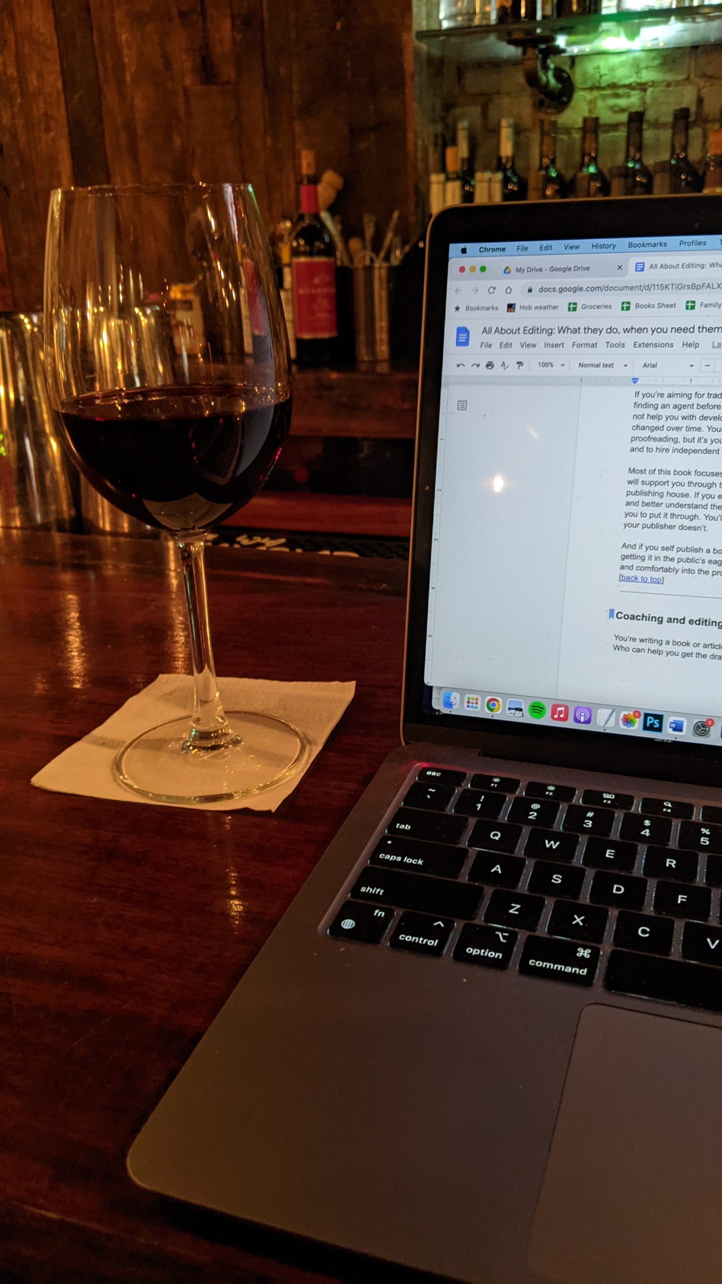 A glass of red wine over a small white paper napkin, next to the edge of a laptop, on a dark wood bar. Wine bottles and lights are out of focus in the background.
